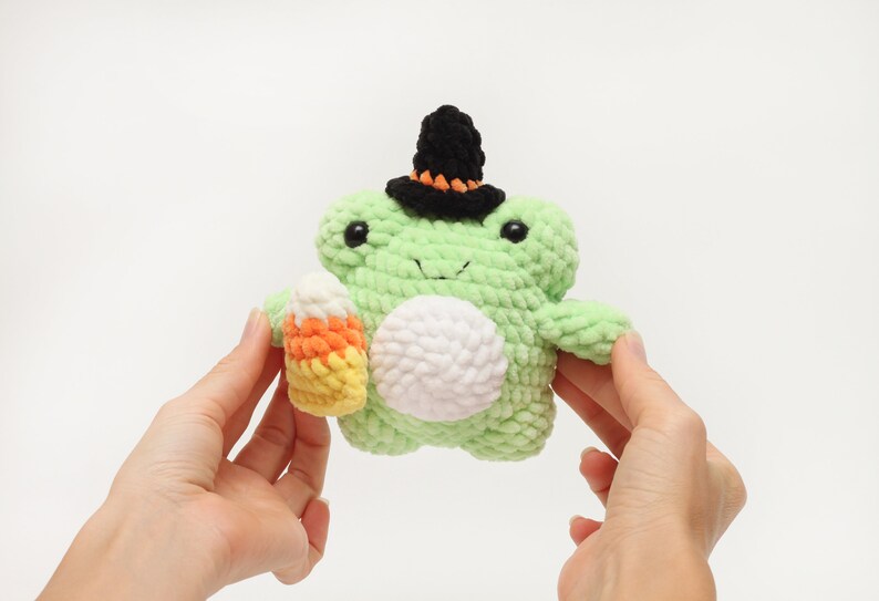May include: A handmade, light green crochet frog wearing a black witch hat with orange trim. The frog holds a candy corn in its hand. The frog has a white belly and black eyes. A Halloween-themed plush toy.