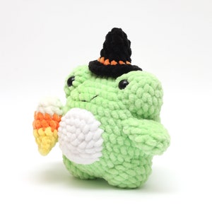 May include: A handmade, light green crochet frog plushie wearing a black witch hat with orange trim. The frog holds a candy corn in its hand. The plushie has a white belly and black eyes, perfect for Halloween.