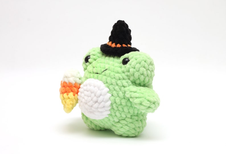 May include: A handmade, light green crochet frog plushie wearing a black witch hat with orange trim. The frog holds a candy corn in its hand. The plushie has a white belly and black eyes, perfect for Halloween.
