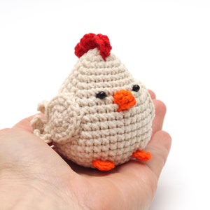K&ouml;nnte beinhalten: Ein geh&auml;keltes wei&szlig;es H&uuml;hner-Spielzeug mit einem roten Kamm, einem orangefarbenen Schnabel und schwarzen Augen. Das Huhn sitzt in der Handfl&auml;che.
