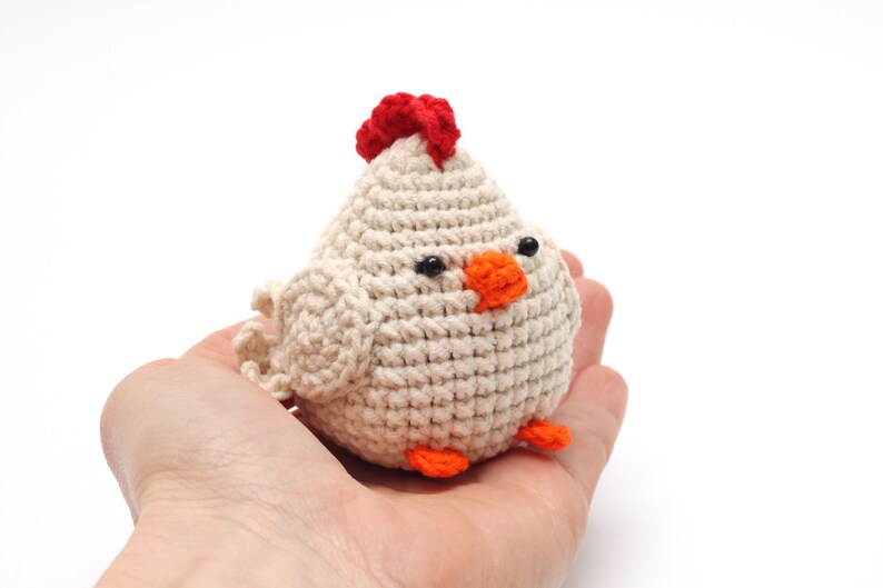 K&ouml;nnte beinhalten: Ein geh&auml;keltes wei&szlig;es H&uuml;hner-Spielzeug mit einem roten Kamm, einem orangefarbenen Schnabel und schwarzen Augen. Das Huhn sitzt in der Handfl&auml;che.