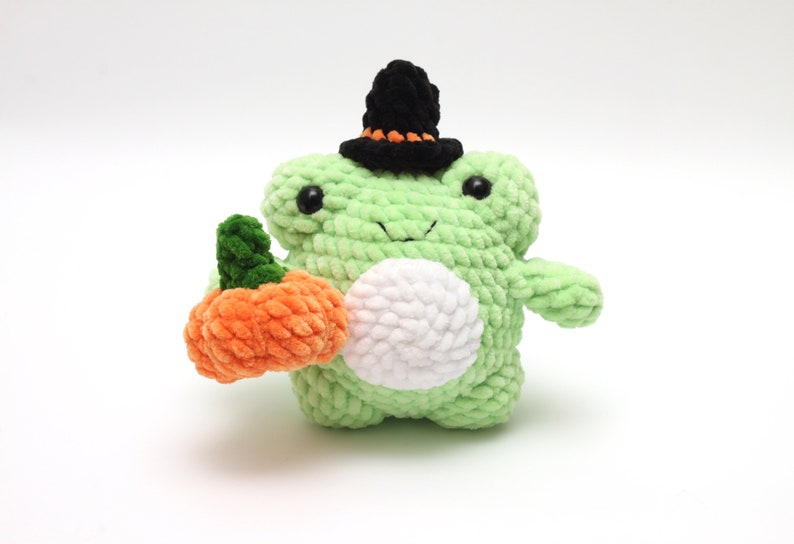 May include: A green crocheted frog wearing a black witch hat and holding an orange crocheted pumpkin.