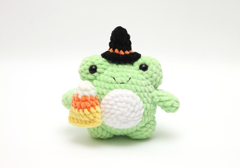 May include: A green crocheted frog wearing a black witch hat and holding a crocheted candy corn.