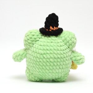 May include: A handmade, light green, plush creature with a black witch hat featuring orange trim. The stuffed animal is holding a small bundle of candy corn. The toy is made of soft, textured yarn.