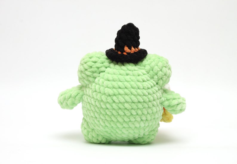 May include: A handmade, light green, plush creature with a black witch hat featuring orange trim. The stuffed animal is holding a small bundle of candy corn. The toy is made of soft, textured yarn.