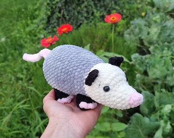 May include: A crocheted plush toy shaped like an opossum. The opossum is gray and white with black eyes and a pink nose. It has a long, pink tail.