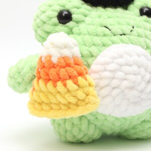 May include: A light green crocheted frog plushie with black eyes and a small black hat. The frog is holding a crocheted candy corn in yellow, orange, and white. The plushie has a white belly.