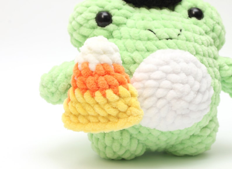 May include: A light green crocheted frog plushie with black eyes and a small black hat. The frog is holding a crocheted candy corn in yellow, orange, and white. The plushie has a white belly.
