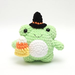 May include: A handmade green crochet frog wearing a black witch hat with orange trim. The frog holds a candy corn treat. The frog has black eyes and a white belly. A Halloween-themed plushie.