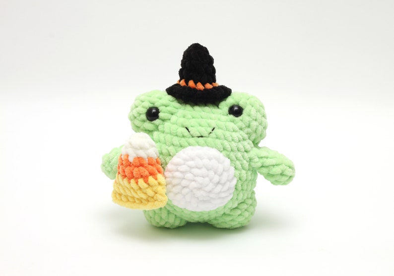 May include: A handmade green crochet frog wearing a black witch hat with orange trim. The frog holds a candy corn treat. The frog has black eyes and a white belly. A Halloween-themed plushie.