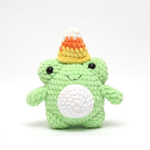 May include: A handmade green crochet frog plushie with black safety eyes and a white belly. The frog is wearing a candy corn hat with yellow, orange, and white stripes. A cute, whimsical Halloween decoration.
