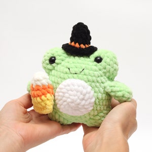 May include: A handmade, light green crochet frog plushie wearing a black witch hat with orange trim. The frog holds a candy corn in its hand. The frog has a white belly and black eyes. A Halloween-themed item.
