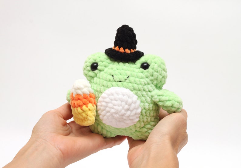 May include: A handmade, light green crochet frog plushie wearing a black witch hat with orange trim. The frog holds a candy corn in its hand. The frog has a white belly and black eyes. A Halloween-themed item.