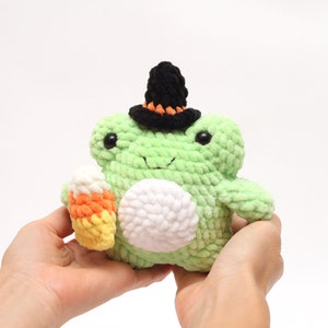 May include: A green crocheted frog wearing a black witch hat with orange trim and holding a candy corn.