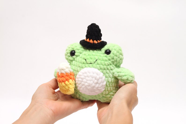 May include: A green crocheted frog wearing a black witch hat with orange trim and holding a candy corn.