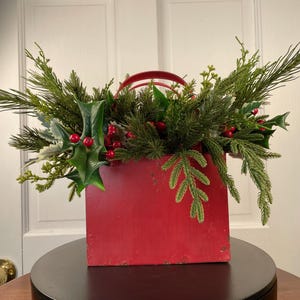 May include: A red wooden box with a red handle, filled with faux greenery, holly leaves, and red berries. The arrangement is a festive holiday decoration, perfect for seasonal displays. The box is approximately 8 inches wide.