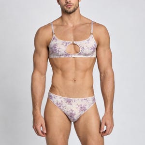 May include: A two-piece lingerie set featuring a light-colored fabric with a purple floral pattern. The top is a bralette with thin adjustable straps and a circular cutout at the center chest. The bottom is a matching pair of high-cut briefs.