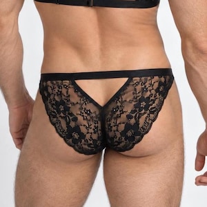 May include: Black lace underwear featuring a floral pattern with a scalloped edge and a horizontal strap across the back. The garment is constructed with sheer black lace fabric and includes a solid black elastic waistband and trim details.