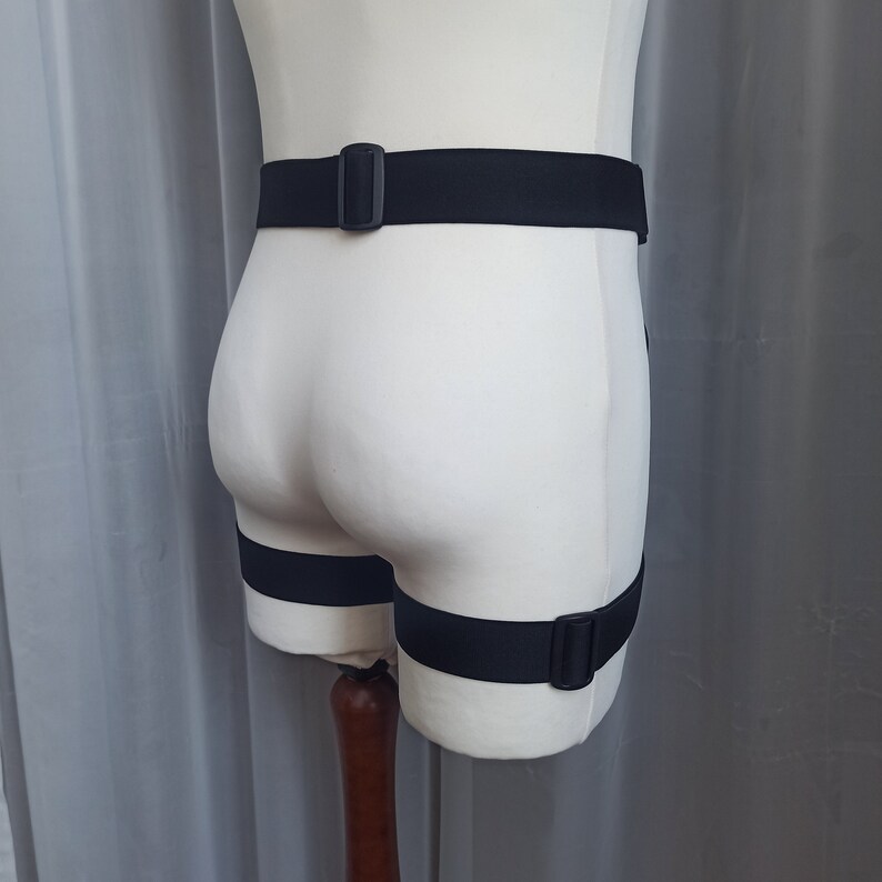 K&ouml;nnte beinhalten: Schwarze verstellbare Riemen mit Schnallen, getragen an einem wei&szlig;en Mannequin. Die Riemen werden um die Taille und die Oberschenkel getragen.