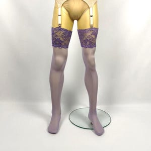 Puede incluir: Medias transparentes de color lavanda hasta el muslo con bandas de encaje morado ancho e intrincado en la parte superior. Las medias están sujetas a un liguero beige con tiras blancas ajustables y clips metálicos, sobre un maniquí.