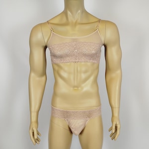 Puede incluir: Conjunto de lencería color nude sobre un maniquí. El conjunto incluye un bralette con detalles de encaje y tirantes ajustables, y un tanga a juego. El bralette tiene una parte superior de malla transparente. La lencería se muestra sobre un fondo liso.