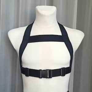May include: A black adjustable harness with a buckle closure. The harness has two straps that go over the shoulders and a strap that goes across the chest.