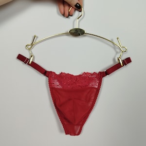 Könnte beinhalten: Roter String-Tanga mit Spitzendetails, aufgehängt an einem goldfarbenen Kleiderbügel. Der String hat verstellbare Träger und einen zarten, durchsichtigen Stoff. Der Kleiderbügel hat eine dekorative ovale Mitte.