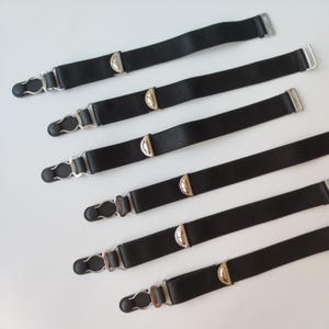 Può includere: Sei giarrettiere elastiche nere con fibbie in metallo argentato e dettagli decorativi. Le giarrettiere sono disposte in fila su una superficie bianca.