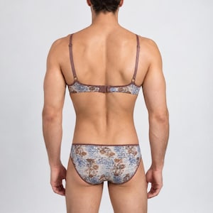 May include: A matching lingerie set consisting of a bra and bikini-style underwear. Both pieces feature a light-colored fabric with a detailed print of brown floral motifs and blue cursive script text, finished with solid brown trim and adjustable straps.