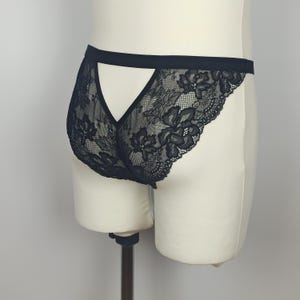 Pode incluir: Calcinha de renda preta com um padrão floral. A roupa íntima apresenta um design recortado nas costas e uma faixa preta na cintura. A renda delicada cria um efeito transparente.