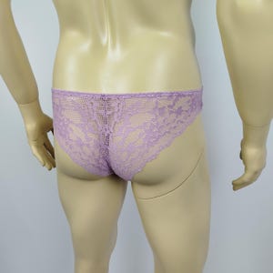 Puede incluir: Ropa interior de encaje lavanda. La delicada lencería presenta un patrón de encaje floral. La ropa interior es de color morado claro y tiene un corte de cintura baja. La prenda se exhibe en un maniquí.