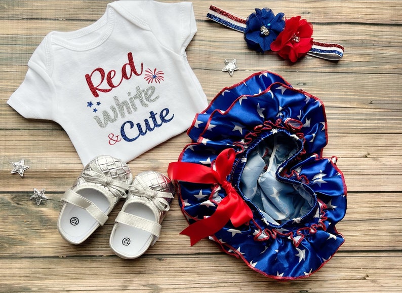 May include: A baby outfit set in red, white, and blue with a white bodysuit that says "Red white & Cute" with silver glitter accents, a blue and white star print ruffle bloomers with a red bow, and silver shoes.