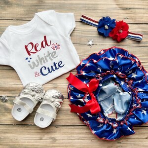 May include: A baby outfit set in red, white, and blue with a white bodysuit that says "Red White & Cute" with a firework graphic, a blue and white star print ruffle skirt with a red bow, and a red, white, and blue headband with a red and blue flower.