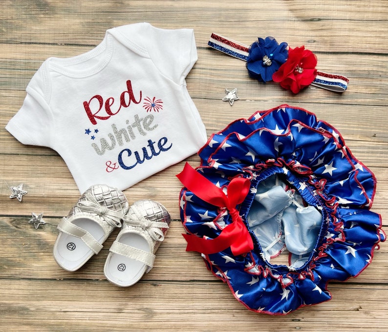 May include: A baby outfit set in red, white, and blue with a white bodysuit that says "Red White & Cute" with a firework graphic, a blue and white star print ruffle skirt with a red bow, and a red, white, and blue headband with a red and blue flower.