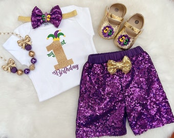 May include: A white onesie with gold glitter 'my 1st birthday' text and a purple and green jester hat. A purple sequined skirt with a gold bow. Gold shoes with purple and green flower embellishments.