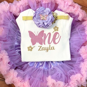 May include: A white onesie with a pink glitter butterfly and the text "One" in pink glitter and "Zayla" in gold glitter. The onesie is paired with a lavender tulle tutu with pink ruffles and a lavender flower headband with a pink butterfly.