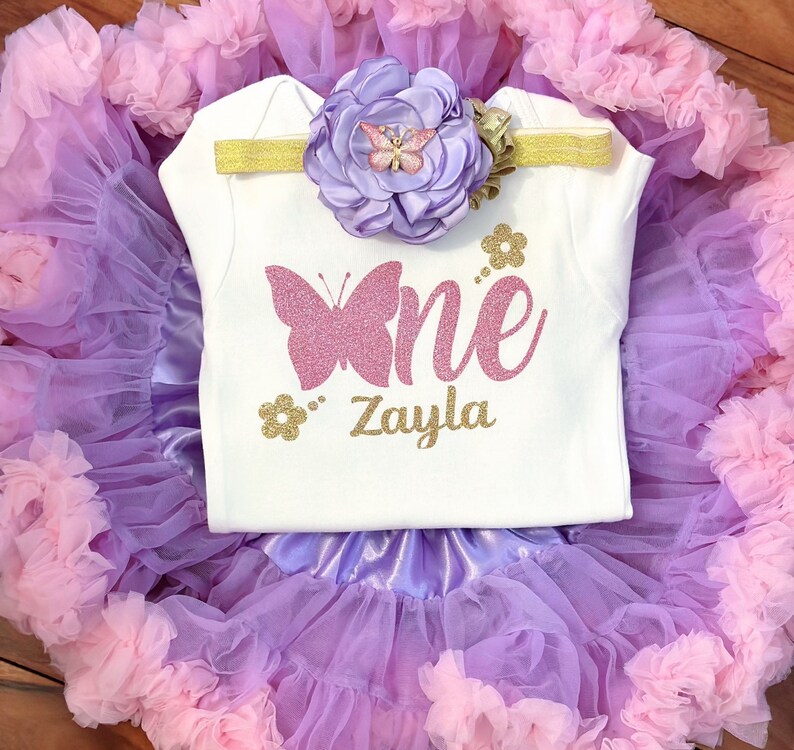 May include: A white onesie with a pink glitter butterfly and the text "One" in pink glitter and "Zayla" in gold glitter. The onesie is paired with a lavender tulle tutu with pink ruffles and a lavender flower headband with a pink butterfly.