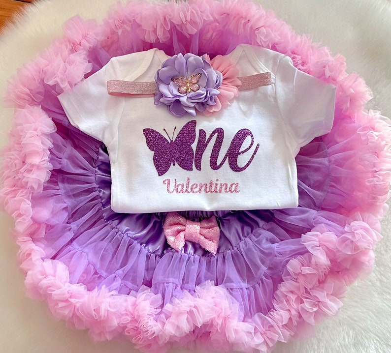 May include: A purple and pink tutu with a white onesie that says "one" with a purple glitter butterfly and the name "Valentina" underneath. The onesie has a pink and purple flower headband.