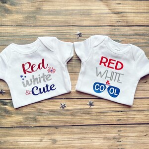May include: Two white baby onesies with red, white, and blue text. One onesie says "Red white & Cute" with a firework graphic. The other onesie says "Red white & Cool" with a graphic of sunglasses.