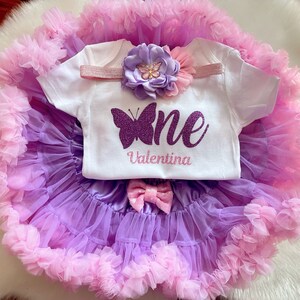 May include: A purple and pink tulle tutu skirt with a white onesie that says "one" with a purple glitter butterfly and the name "Valentina". The onesie also has a pink headband with a purple flower.