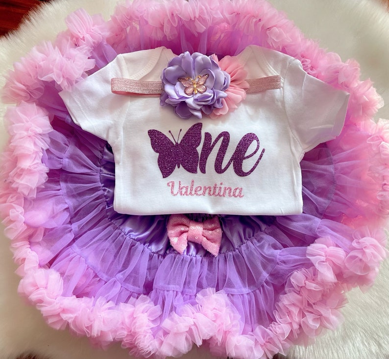 May include: A purple and pink tulle tutu skirt with a white onesie that says "one" with a purple glitter butterfly and the name "Valentina". The onesie also has a pink headband with a purple flower.