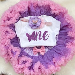 May include: A white onesie with a purple butterfly and the word "one" in purple glitter. The onesie is paired with a purple and pink tulle tutu and a headband with a purple flower and glitter.