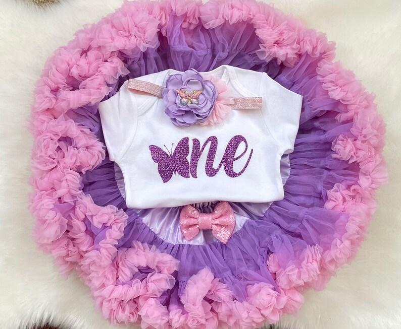 May include: A white onesie with a purple butterfly and the word "one" in purple glitter. The onesie is paired with a purple and pink tulle tutu and a headband with a purple flower and glitter.