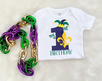 May include: A white babygrow with a purple number one, a gold fleur-de-lis, and the text "my 1st birthday". The babygrow is decorated with a green and gold jester hat. A purple, green, and gold chain is in the background.