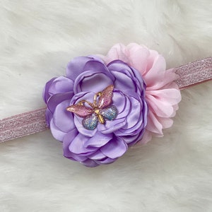 May include: A purple and pink flower headband with a sparkly gold butterfly accent. The headband is made of elastic and is perfect for a baby or toddler.