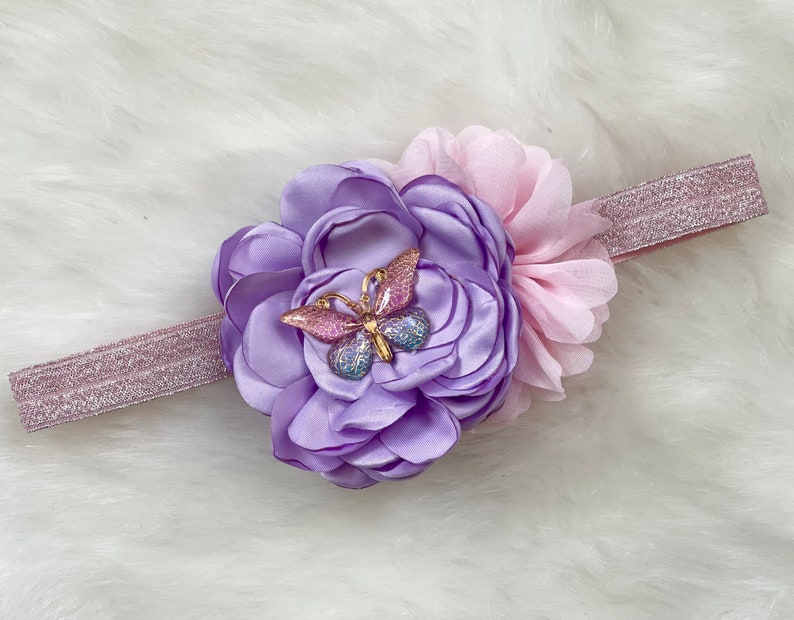 May include: A purple and pink flower headband with a sparkly gold butterfly accent. The headband is made of elastic and is perfect for a baby or toddler.