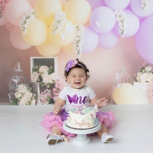 May include: A baby girl wearing a purple tutu and a white shirt with a purple butterfly and the word "one" on it, smiles while sitting in front of a cake decorated with butterflies. The cake is on a white cake stand. The background is a pink wall with purple and yellow balloons and white flowers.