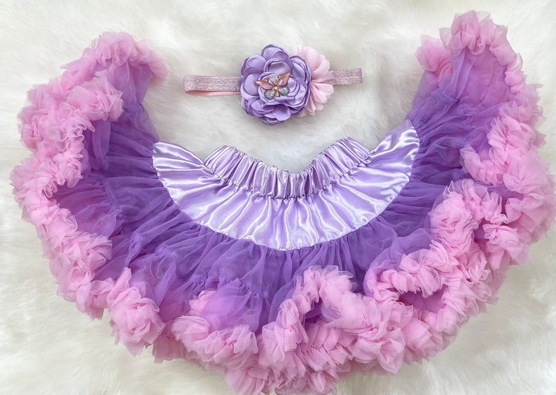 May include: A purple and pink tulle tutu skirt with a satin waistband and a matching flower headband with a butterfly embellishment.