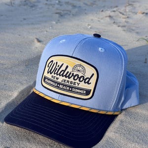 May include: A light blue baseball cap with a dark navy blue brim and a yellow rope detail. The front features a rectangular patch with the text Wildwood New Jersey Boardwalk Beach Summer and a graphic of a Ferris wheel in black and yellow.