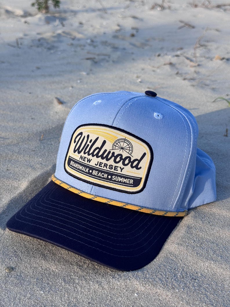 May include: A light blue baseball cap with a dark navy blue brim and a yellow rope detail. The front features a rectangular patch with the text Wildwood New Jersey Boardwalk Beach Summer and a graphic of a Ferris wheel in black and yellow.